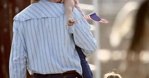 A child sits on someone's shoulders holding a small American flag.