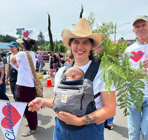 A woman wearing a straw hat and baby carrier holds a small flag and a fern branch outdoors.