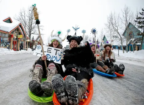 People in costume ride sleds down a snowy street.