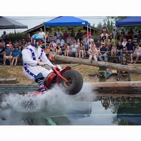 A person dressed as a clown rides a three-wheeled motorcycle into a pool of water in front of a crowd.