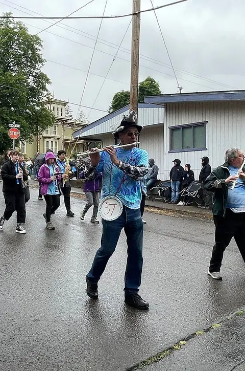 A street performer plays a flute in a procession while wearing a tie-dye shirt and hat.