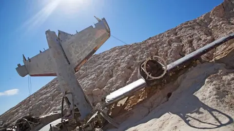 An airplane wreckage rests on a hillside under a bright sun.