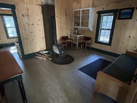An interior view of a rustic cabin with wood-paneled walls, a wood stove, and simple furniture.