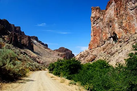 A dirt road winds through a canyon with rocky cliffs and green bushes under a blue sky.