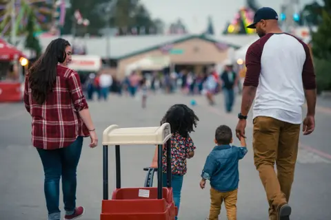 A family walks together at an outdoor event, pulling a red wagon.