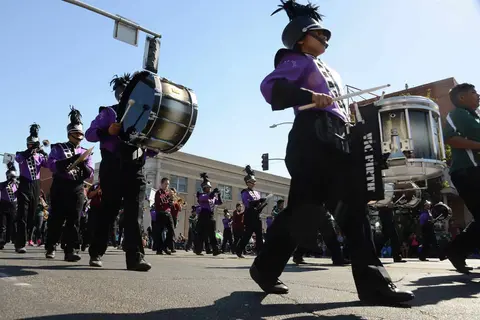 A marching band with percussionists and musicians walks down a street.
