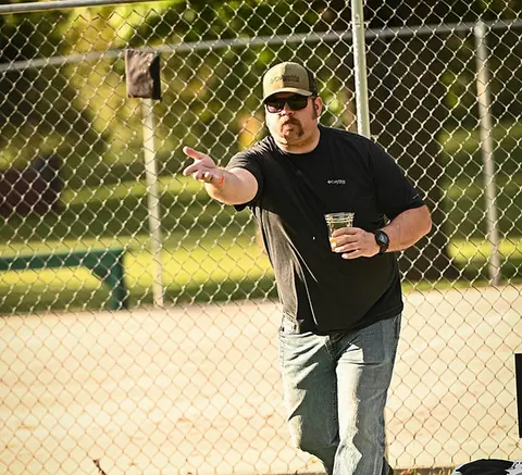 A man throws an object while holding a cup in front of a chain-link fence.