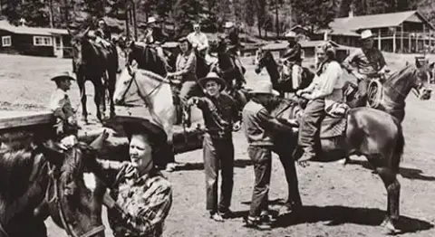 A historic black and white photograph shows a group of people on horseback outdoors.