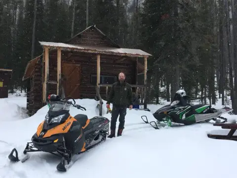 A person stands in the snow in front of a rustic cabin with two snowmobiles parked nearby.