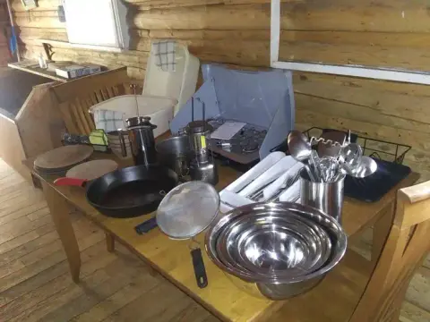 A wooden table inside a rustic cabin holds camping gear, cookware, and utensils.