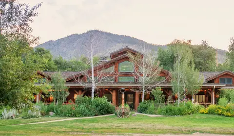A large wooden lodge building set against a mountain backdrop with lush landscaping.