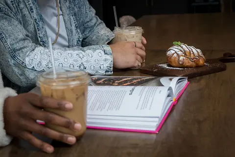 Two people sitting at a wooden table with iced coffee, a book, and a pastry.
