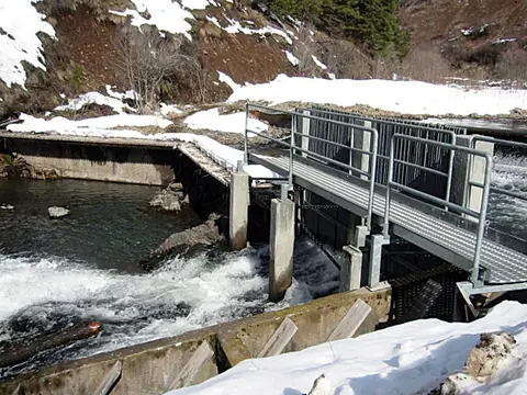A small dam and metal walkway over a rushing stream with snow on the banks.