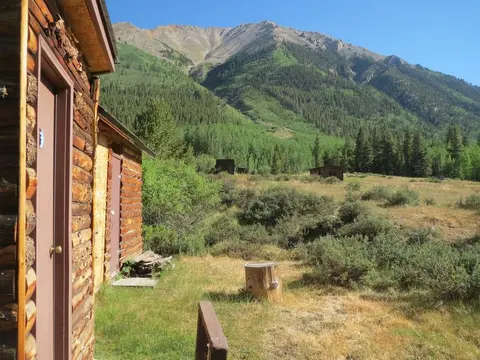 A rustic log cabin sits in a grassy valley with a large mountain in the background.