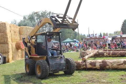 A person operates a skid steer loader outdoors near stacked hay bales and a crowd.