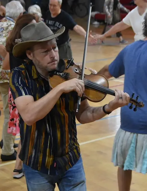 A man wearing a hat plays a violin indoors while people dance in the background.