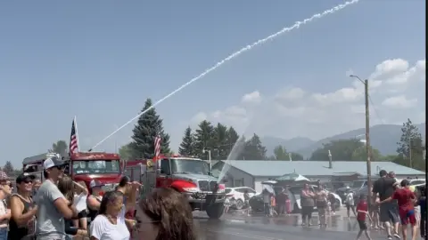 A crowd watches fire trucks spray water in the air during an outdoor event.