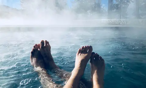 Two pairs of feet relax in a steaming outdoor pool surrounded by trees.