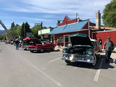 Classic cars are parked along a street in front of a red building with people walking around.
