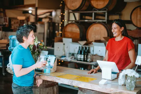 A woman in a blue shirt holds a glass and talks to a woman behind a counter in a wine tasting room.
