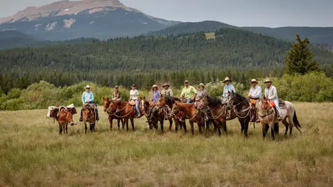 A group of riders on horseback in a grassy field with mountains in the background.