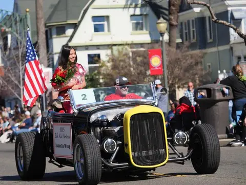 A young woman holding flowers sits in a vintage car in a parade.