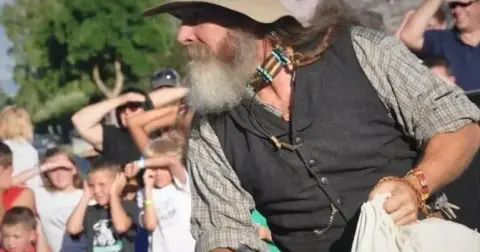 A bearded man in historical costume holding a white pouch stands before a crowd of spectators.