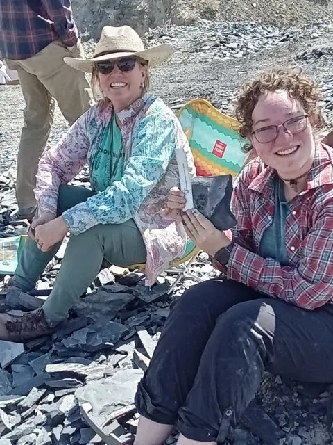 Two women sitting outdoors on rocky ground holding a fossil specimen.
