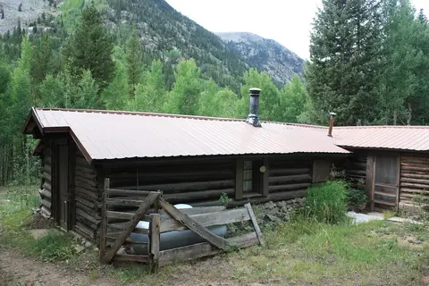 A log cabin sits in a clearing surrounded by trees and mountains.