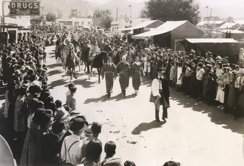 A historic black and white photograph showing a large crowd gathered for a parade or street event.