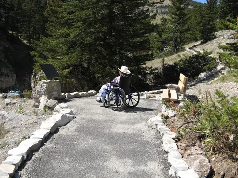 A person in a wheelchair on a paved path in a mountainous landscape.