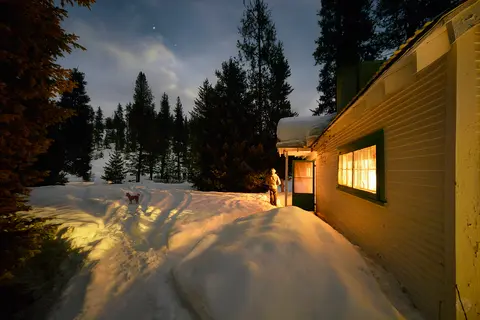 A glowing cabin stands in a snowy landscape at night.