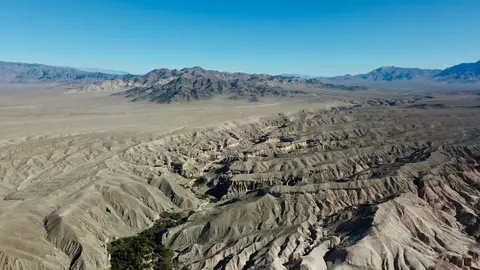 Aerial view of an arid desert landscape with eroded hills and mountains under a blue sky.