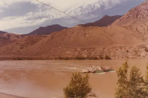 A ferry crosses a wide river with arid hills in the background.