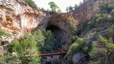 A metal footbridge crosses a rocky gorge in front of a large cave opening.