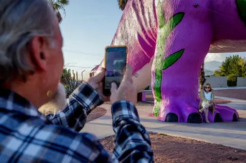 A man takes a photo of a woman sitting on the foot of a large dinosaur statue.