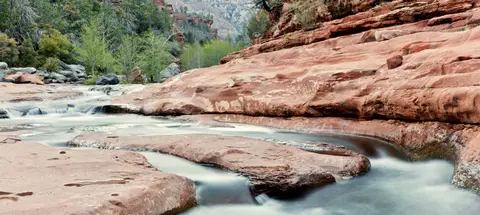 A stream flows over smooth red rock formations surrounded by trees and mountains.