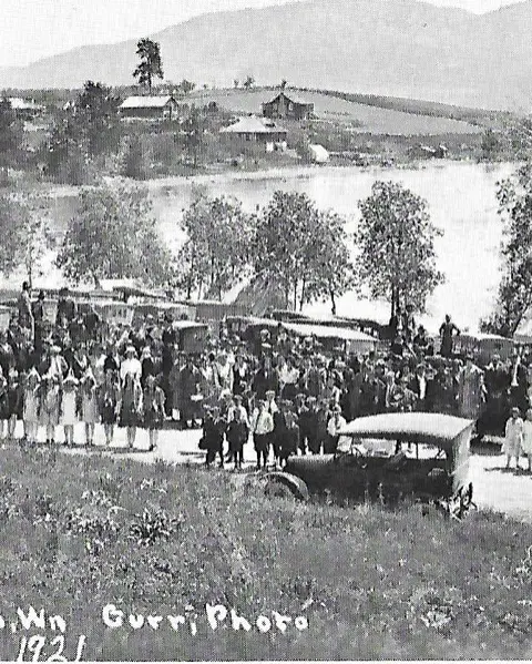 A historic black and white photograph showing a large crowd gathered outdoors near water.