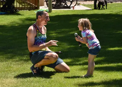 A person with brightly colored hair kneels on the grass while talking to a child holding a microphone.