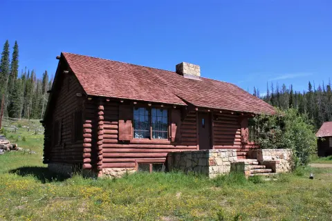 A log cabin exterior sits in a grassy, forested area under a clear blue sky.