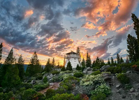 A white lookout tower stands on a hill surrounded by trees under a colorful sunset sky.