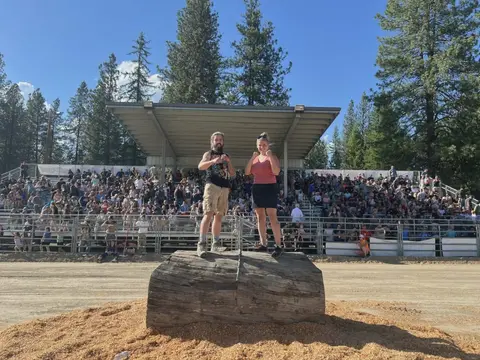 Two people pose on a large wooden log in front of a grandstand filled with a crowd.