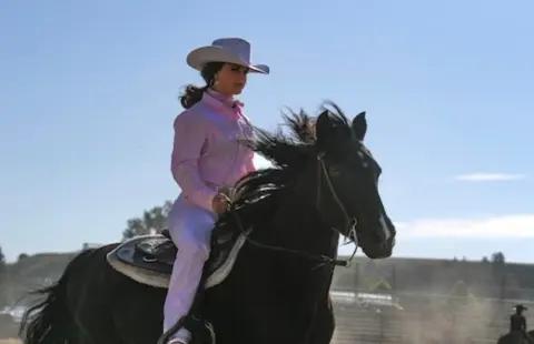 A woman in a cowboy hat and pink shirt rides a black horse outdoors.