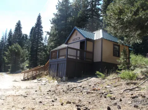 A small cabin stands among pine trees in a mountain setting.