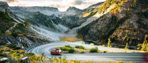 A red van drives along a winding mountain road with rocky peaks in the background.