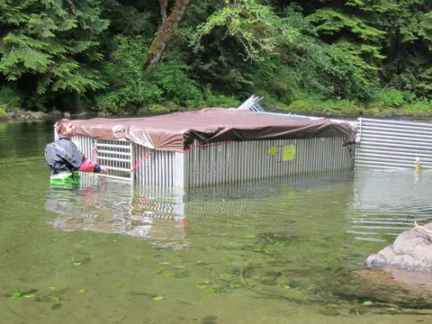 A person in a wetsuit stands in a river next to a submerged metal enclosure covered with a tarp.