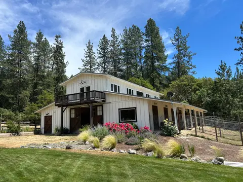 A white barn with a second-story balcony stands surrounded by trees and landscaping under a blue sky.