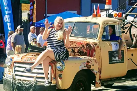 A woman waves while sitting on the hood of an old truck during an outdoor event.