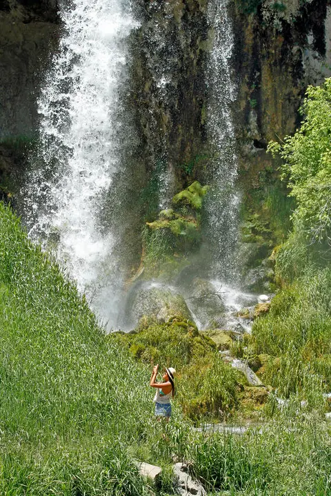A person taking photos of a waterfall amidst lush greenery.