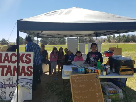 A family gathers under a canopy tent with a food stand setup outdoors.